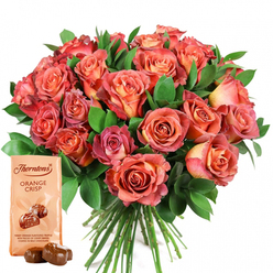 There's a special energy in this hand-crafted gift set, reminiscent of Clerkenwell's spirited charm and creative pulse. A striking bouquet of roses commands attention, the blooms softly illuminated and bursting with layered hues-from rich coral and warming peach to the faintest blush of cream, each flower curling into itself with a kind of gentle movement. The roses' petals offer a tactile richness, velvety and inviting, as their ombre transitions create an impression of twilight falling over Exmouth Market. Lush, deep green foliage-most likely eucalyptus or salal-cradles the flowers in a fresh embrace, accentuating the bouquet's fullness while hinting at the local parks around Spa Fields. Setting off this floral centrepiece is a pouch of Thorntons Orange Crisp, light beige in colour, positioned carefully in the lower part of the composition and showing off the iconic logo with its illustration of glossy, rounded truffles. In front, two unwrapped truffles rest temptingly, promising a zesty orange interior encased in smooth milk chocolate-a fragrant and luxurious treat. It's a composition that appeals to all the senses: you can almost feel the coolness of the greenery, the silkiness of the petals, and the crisp snap of chocolate. Thoughtfully assembled, this Clerkenwell-inspired gift conjures up feelings of romance, gratitude, and celebration-suiting everything from Mother's Day to spontaneous gestures of affection for someone special.