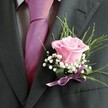 There's poetry woven into the folds of this boutonniere-a composition carefully arranged in the bustling flower shops of Clerkenwell, near Smithfield's markets. At its centre, a blush-pink rose opens quietly, its velvet petals catching soft light and whispering of beginnings, love, and gentle optimism. The delicate fragrance-imagined or perhaps remembered from past spring mornings-hovers over snowy tufts of baby's breath, lending an airy softness akin to morning clouds rising over the rooftops of Clerkenwell Green. Behind, slender tendrils of green foliage and bold, dark leaves create a backdrop of subtle forest freshness, their curves bringing flow and movement to the design. Nestled at the boutonniere's base, a silky lavender ribbon gathers all elements together, its sheen glimmering playfully as the wearer moves. The flowers are pinned neatly on the lapel of a charcoal-grey suit jacket, the jacket's textured weave and crisply stitched buttonhole revealing the careful preparation behind the scene. Beside it, a fuchsia pink tie makes a joyful statement against a pale grey dress shirt, the whole ensemble glowing in soft indoor light. Each component is selected by a seasoned florist to embody the anticipation and unity of special occasions-whether a wedding at one of Clerkenwell's unique venues or a personal milestone worth celebrating amidst London's vibrant urban heart.