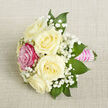 Laid diagonally across a pale beige, finely woven fabric that suggests a relaxed yet polished setting, this petite bouquet feels like a moment of romance captured in still life, just as you might find in a Clerkenwell townhouse before guests arrive. Five ivory-white roses form the core of the arrangement, each bloom opening generously, their layered petals soft and rounded, giving the piece a sense of fullness despite its compact size. Off to the front-left, a single fuchsia pink rose steps forward, its saturated colour providing a lively burst that instantly catches the gaze and adds character, much like an unexpected splash of street art along nearby Clerkenwell Road. Wisps of baby's breath weave through the roses, their tiny white florets clustered like delicate froth, creating a light, cloud-like veil that softens the composition. Around the flowers, deep green pointed leaves rise and curve, offering structure, contrast, and a hint of natural woodland texture. The gathered stems are smoothly wrapped in a bright pink satin ribbon, forming a slim, polished handle that feels both comfortable to hold and elegantly decorative. Layered over this, aligned rows of small, luminous white pearls reflect the ambient light, suggesting thoughtful attention typical of an experienced local florist preparing for weddings, proms, or anniversaries across Clerkenwell and nearby Farringdon. Photographed from a slightly raised, close perspective with gentle, even lighting, every detail stands clear: the plush petals, the airy gypsophila, the gleam of satin and pearls. The bouquet's overall impression is one of intimate celebration and gentle sophistication, ideal as a corsage, boutonnière, or petite centerpiece for a quietly memorable occasion.