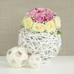 A stylish floral centerpiece is captured in soft, even light, showcasing a domed bouquet of roses perched on a distinctive white wicker globe, ideal for a refined celebration in Clerkenwell. The arrangement is composed of around a dozen fresh roses, closely clustered into a rounded, almost cushion-like form. About half of the blooms are a gentle pastel pink, touching on lavender or fuchsia tones, while the others are a creamy, buttery yellow softly fading to ivory at the edges. Each rose reveals different stages of opening, from tight, hopeful buds to fully unfurled blossoms with velvety, layered petals, giving a sense of richness without feeling overwhelming. Threaded between the roses are airy sprigs of white baby's breath, their tiny blossoms creating soft, cloud-like accents that lighten the composition, reminiscent of the delicate spring blossoms you might glimpse on a walk from Clerkenwell Green towards Rosebery Avenue. The flowers are arranged in a spherical vase made from thin white-painted branches or wicker, delicately intertwined to form an open, lattice-style structure that looks both handcrafted and contemporary. Resting on the pristine white surface beside it are two smaller decorative orbs made of natural light wood strips, echoing the woven texture of the vase; one orb is larger, the other smaller, adding gentle asymmetry and depth. Behind, a neutral beige or light taupe wall with a subtle fabric-like texture offers a warm, unobtrusive backdrop that allows the soft pinks, creams, and crisp whites to stand out. The overall effect is serene, romantic, and thoughtfully arranged, suited to a wedding table, anniversary dinner, or elegant home in the heart of Clerkenwell.