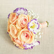 Captured from a gentle overhead angle on a neutral, woven-beige backdrop, this bouquet is an ode to soft colour contrasts and delicate textures. The main structure is formed by cream-tinged apricot-peach roses, their petals unfolding in concentric layers that offer both volume and a plush, inviting surface. Interspersed among them are ruffled lisianthus flowers, white at the core with a distinctive violet-purple edging that frames each bloom like a fine brushstroke; glimpses of yellow stamens provide tiny, bright focal points within those cool-toned petals. Scattered throughout are feathery sprays of baby's breath - minute white blossoms that breathe lightness into the composition and prevent the arrangement from feeling heavy. The stem-wrap is a careful study in refinement: a soft peach ribbon envelops the handle, studded lengthwise with rows of small pearlescent beads, creating a sensual tactile detail meant to feel as luxurious in hand as it looks in photos. The exposed stem ends are trimmed clean and reveal fresh green hues that anchor the bouquet visually. Under diffuse daylight the colours read true and tender, and the textures - from velvety rose petals to the papery lisianthus edges and whisper-fine Gypsophila - are clearly legible. This bouquet embodies a serene romanticism suited to ceremonies in Clerkenwell chapels or intimate receptions near the Leather Market, offering an impression of thoughtful floristry and understated celebration.