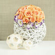 An elegant dome of roughly two to three dozen apricot and soft peach roses forms the heart of this arrangement, each blossom layered with velvety petals-some opened wide, others curled tight-creating a richly textured silhouette. Encircling the rose dome, white lisianthus blooms punctuate the warmth with cream petals brushed at the edges in a dramatic deep violet, lending a painterly, slightly edgy detail. Interspersed throughout are tiny clusters of white gypsophila, their feathery spray adding air and a whisper of classic bridal sweetness. The bouquet is set in a contemporary rounded white woven orb, an airy lattice of reeds or wicker that feels both sculptural and handwoven; its bright surface picks up soft studio light and contrasts the flowers' warm blush. Arranged to the left on the same white surface are two open-woven decorative spheres in pale wood or bamboo, echoing the basket's texture and enhancing the balanced, natural aesthetic. The backdrop is a muted linen beige, gently out of focus, allowing the floral forms to remain the clear focal point. As a Clerkenwell florist I imagine this piece on a table near St John Street or sent to a celebration hosted close to Farringdon: it reads as intimate, refined, and quietly joyful. This composition-prepared by Clerkenwell Flowers with an eye for texture and seasonality-suggests a soft, honeyed rose scent and an atmosphere of thoughtful elegance, perfect for weddings, anniversary gifts, or a polished home centerpiece.