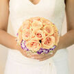 This bridal arrangement reads like a soft poem-apricot roses gathered into a symmetrical, sunlit cluster, each rose unfurling with a creamy, satin-like texture that captures and reflects a warm, peachy glow. Encircling the roses is a neat ring of purple-tipped lisianthus, the cool mauve edges recalling twilight clouds over Clerkenwell Green and offering an elegant, almost painterly contrast to the roses' warmth. A delicate lattice of white baby's breath weaves through the bouquet, introducing a weightless lilt that makes the composition feel airborn, as if a summer breeze might lift it. The petals show delicate veins and subtle shading; light falling from one side reveals their layered curvature and the soft shadows between blooms. Holding this bouquet against bridal lace or a simple satin gown, you notice the scent first-clean, sweet rose tempered by leafy freshness-then the reassuring craftsmanship of every tie and stem. As a local florist who knows St John Street and the markets around Farringdon, I picture it delivered with care from our Clerkenwell Flowers workshop, fresh and ready for photographs in the morning light. The overall mood is timeless and refined: lush yet restrained, intended to be the quiet centrepiece of vows exchanged beneath old brick facades. It feels like a tradition renewed-poised, joyful, and designed to be held close throughout a life of ordinary, treasured moments.
