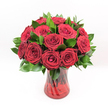 A studio photograph shows a classic bouquet of deep red roses arranged in a rich ruby glass vase, set against a pure white background so that every petal and leaf stands out with clarity. Around a dozen or more velvety crimson blooms are gathered tightly together, their centres still gently furled, creating that perfect, romantic cup shape associated with timeless love. The roses form a rounded, symmetrical dome, each flower turned outward as if a careful florist has positioned them one by one, making sure no bloom is hidden. Threaded between the blossoms are glossy, dark green leaves, likely Ruscus, their pointed tips and waxy sheen adding contrast and structure, and giving the arrangement a full, luxurious profile. The vase itself is a translucent ruby red, with a subtle ribbed or swirling texture that catches the light and echoes the intensity of the petals above. Through the coloured glass, the upright stems appear as darker, shadowy lines, adding depth and a sense of natural movement. The lighting is bright, crisp, and even, typical of a professional florist's product photograph, highlighting the freshness of the flowers, the smooth glass, and the lush greenery. The overall feeling is one of romance and passion, suitable for anniversaries, Valentine's Day, or a dramatic declaration of love. It is the sort of bouquet a local Clerkenwell resident might send from a favourite flower shop for delivery to a townhouse off Exmouth Market or a smart apartment near Farringdon, confident that this classic display of red roses will say everything that words cannot.