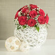 At the heart of this composition is a lush, perfectly rounded bouquet of red roses, gathered so closely that their blooms seem to melt into one another, forming a rich, velvety crown of colour. Each rose is at full, symmetrical openness, the petals tightly yet elegantly coiled, giving that sense of just-unfolded freshness you might expect from a same-day delivery in Clerkenwell. Around and between the scarlet blossoms float delicate tufts of white baby's breath, their tiny flowers like flecks of soft foam against the deep red sea of petals. The contrast between the bold roses and the airy gypsophila adds a lightness that feels ideal for wedding centrepieces or intimate celebrations in nearby Farringdon and around St John Street. A few deep green leaves emerge discreetly, lending a natural depth and hinting at the stems hidden within the vessel. The bouquet rises from a round, snow-white vase created from interwoven strips that resemble fine wicker or rattan, curved into a hollow sphere. This open lattice structure allows slivers of light to pass through, casting subtle shadows and giving the entire arrangement an architectural flair. On the white tabletop beside it sit two decorative woven orbs in a lighter, creamy tone, repeating the pattern and tying the scene together with understated style. Behind everything, a neutral beige-grey wall provides a quiet backdrop, ensuring that the roses and intricate textures retain full visual attention. The soft, diffuse lighting evokes a calm afternoon, making the display feel both refined and inviting, like a thoughtfully styled corner of a Clerkenwell apartment prepared for a romantic evening.
