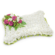 A plush, rectangular memorial pillow of blooms sits against a clean white backdrop, its surface entirely covered with a tightly packed carpet of small, pristine white chrysanthemums that read like soft fabric to the eye and imagine the touch. The cushion-like form, with gently rounded corners, carries a sense of calm and repose; the chrysanthemums' tiny petals create a uniform, cloud-like texture that suggests peace and timeless rest. Anchoring the composition is an elegant spray set into the upper left corner: several deep magenta-to-fuchsia roses open in layered spirals, their velvety petals contrasting warmly with the cool white field. Nestled among the roses are delicate white freesias and tiny white buds that add fragrance and a whisper of brightness, while slender fern fronds and glossy green leaves weave through the cluster, lending depth and a natural frame. Along the pillow's edge a pale green satin ribbon has been pleated into a discreet looped border that peeks out beneath the chrysanthemum surface, softening the silhouette and introducing a hint of fresh colour. Soft, even lighting casts a gentle shadow beneath the arrangement, emphasizing its three-dimensional form without distraction. This composition reads as a traditional sympathy tribute-quiet, reverent and crafted with exacting care-a floral pillow chosen for funerals or graveside remembrance where the intention is to convey comfort, enduring memory and a restful farewell. As a local touch, imagine this piece arriving for a service near Exmouth Market or the quiet squares around Clerkenwell Green, a simple, dignified expression of condolence suited to the neighbourhood's thoughtful pace.