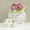 This image captures a refined floral centerpiece that could easily sit on a wedding table in a Clerkenwell townhouse near Myddelton Square, bringing a sense of quiet romance to the setting. At its core is a low, perfectly rounded bouquet of roses in shades of soft pink and twilight lavender, each bloom fully open so that the spiral of petals and the slightly darker pink edges are clearly visible. The roses are interwoven with bright green button chrysanthemums whose small, domed flowers provide a lively texture, like tiny cushions of fresh moss tucked among the petals. Around the bottom edge, clusters of petite white filler flowers create a gentle halo, their delicate blossoms making the overall shape look relaxed yet carefully composed. The arrangement is gathered into a spherical vase crafted from slender, white rattan-like twigs that twist and loop around one another, leaving airy gaps that allow light to pass through. This open structure gives the vase a sculptural presence, bridging rustic influences with a contemporary feel that suits modern London interiors. Resting on the white tabletop to the left of the vase are two decorative wicker balls in pale, natural wood tones-one noticeably larger, one smaller-echoing the woven pattern and adding a touch of understated styling, as though the table had been thoughtfully laid for a reception. The background is a softly focused beige fabric, suggestive of linen, which adds warmth without distracting from the flowers. Overall, the composition conveys freshness, softness, and handcrafted detail, an ideal visual for a wedding arrangement or special occasion setup in Clerkenwell.