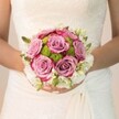 Bathed in the gentle daylight of Clerkenwell's side streets, a bride holds close an elegantly rounded bouquet-a poetic blend of texture and colour. Mauve roses, centre stage, unfurl their plush petals dusted with deeper fuchsia, nodding to romance beneath the intricate, off-white floral lace of her dress. Sprightly lime-green button chrysanthemums are woven throughout, their dewy spheres bursting with vibrant life, recalling the freshness of spring mornings near Farringdon Road. Encircling the heart, dainty white blossoms-possibly freesia or tiny spray roses-add a breath of airy refinement, their subtle scent evoking purity and tender new beginnings. Tucked among the petals, slivers of green buds and leaves impart dimension, while the bride's manicured hands, traceable by blush pink polish and delicate shimmer, cradle each bloom with care. Soft, diffuse lighting reveals every detail, from the delicate gathering of fabric at her waist to the minute veining of every rose. The bouquet's harmonious blend of pastel, green, and white offers an emotional portrait-one of quiet anticipation and love, fitting for weddings held in Clerkenwell's historic halls, and expertly arranged by Clerkenwell's local florists.