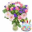 A vibrant, celebratory bouquet sits against a pristine white background, photographed with bright, even light that highlights every petal and leaf. The arrangement is centred on several elegant pink roses, their velvety petals unfurling from pale blush to a warmer mid-pink, lending a classic, romantic core to the display. In front and slightly lower, large pale-pink Gerbera daisies fan their layered, ray-like petals around soft butter-cream centres, adding an airy, cheerful note. Scattered through the massed blooms are clusters of vivid purple filler-likely statice or limonium-whose papery texture and deep hue create lively contrast and visual depth. Small white spray blossoms, perhaps spray carnations or alstroemeria, nestle between the larger flowers to give fullness and delicate highlights, while abundant glossy green foliage frames and supports the composition. The stems sit in a clear glass vase with a gently flared base; the water and green stems are visible, a subtle reassurance of freshness. To the lower right, a small metallic foil balloon shaped like a layered birthday cake provides a playful accent: a white frosted top dotted with colourful sprinkles, a light blue base, and the words Happy Birthday! printed in bright, multi-coloured lettering. Four tiny printed candle motifs in red, yellow, green and blue, each with a small flame, sit atop the cake illustration, reinforcing the celebratory message. The whole picture reads as a thoughtful birthday gift-softly scented, tactile, and well balanced-the kind of cheerful arrangement an independent Clerkenwell florist might place in a shop window near Exmouth Market or Leather Lane to catch passing smiles.