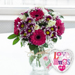 A richly coloured bouquet sits in the centre of the image, as if waiting to be handed to someone special in Clerkenwell after a stroll past Exmouth Market. The arrangement overflows from a classic, rounded clear glass vase placed on a smooth, white surface, the bright light giving everything a crisp, uplifting feel. Several bold gerbera daisies in saturated magenta and fuchsia command attention, their dark central disks lending drama to their cheerful faces. Interspersed with them are clusters of small, decorative chrysanthemums whose petals shift from deep purple at the base to gleaming white tips, each tiny bloom set around a yellow centre that adds playful brightness. Weaving through the more vivid blossoms are creamy white roses, some just beginning to unfurl, contributing a soft, romantic note to the design. Sprays of fine white baby's breath flutter lightly around the stronger colours, creating a misty, lace-like halo that softens the whole bouquet. Fresh green foliage emerges between the flowers, framing them and rounding out the compact, dome-shaped arrangement so typical of a carefully crafted florist design. The glass vase reveals neatly trimmed green stems standing in clear water, while a simple band of rustic twine encircles the neck, tied in a casual bow. From this hangs a small, white, rectangular gift tag printed with a stylised pink rose and easy-going green writing, suggesting something personally chosen rather than mass-produced. To the lower right, partly overlapping the vase, a gleaming silver heart-shaped foil balloon rests, its white centre boldly carrying the phrase Love n' Hugs 24-7 in a fun mix of patterned pink, purple, and blue letters with tiny hearts. The softly blurred, bright background hints at daylight streaming through a nearby window, giving this floral-and-balloon duo the feel of a joyful delivery for a birthday, anniversary, or spontaneous show of affection anywhere around Clerkenwell.
