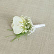 Resting on a softly woven, light beige fabric that recalls the calm interiors of Clerkenwell townhouses, this white orchid boutonnière exudes understated refinement. At its heart sits a single large orchid bloom, petals wide and flawlessly smooth, their pure white surface gently curving around a centre lightly washed with creamy yellow, giving warmth and depth to the flower's serene expression. Clustered closely around the orchid, airy sprays of white baby's breath create a cloud-like frame, each minuscule blossom like a tiny dot of light, balancing the orchid's bold presence with a fine, lace-like texture. Slender strands of vivid green foliage, perhaps fine grass or fern, slip out from beneath the flowers, extending the composition with graceful lines that feel fresh and modern, much like the contemporary galleries and studios tucked around Clerkenwell Road. The stems have been neatly gathered and wrapped in a sheath of white satin ribbon, the fabric smooth and slightly lustrous, forming a comfortable, secure handle ready for pinning onto a lapel. A small satin bow, crafted with care, sits near the top of the wrapping, adding a tailored finish reminiscent of a carefully knotted tie before a ceremony. The boutonnière lies at a gentle angle, and the soft, even lighting reveals the textures-the satin's sheen, the orchid's velvety surface, the tiny blossoms of gypsophila-without harsh shadows. It feels like a moment just before a wedding in one of Clerkenwell's historic venues, a quiet, elegant detail prepared for a groom, best man, or honoured guest who wants a touch of natural grace close to the heart.