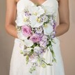 Radiating gentle sophistication, this photograph highlights a bride in Clerkenwell, elegantly holding a voluminous, hand-crafted cascading bouquet against a subtle beige background reminiscent of local sandstone facades. The bouquet's core is formed by pristine white phalaenopsis orchids-petals wide and luminous, centers kissed with yellow-artfully joined by blush pink garden roses that evoke romance and timelessness. Slender stems of lavender-tinted stock and petite white lisianthus, edged with glimpses of purple, spiral through the plush arrangement, lending both layered colour and intricate texture. Wisps of baby's breath and subtle threads of green introduce an ethereal quality and amplify the bouquet's overflowing, teardrop shape. The bride, within her intricately laced strapless gown, draws all focus to the floral composition with a gentle embrace, hinting at the intimacy of a wedding morning near Clerkenwell Green. The mood is one of refined celebration, where floral forms and colours merge seamlessly to honor modern love and historic London charm. This bouquet, as seen here, epitomizes the classic artistry of Clerkenwell florists and is a luminous choice for those seeking wedding flowers that both celebrate personal romance and resonate with the unique spirit of EC1's vibrant streets.