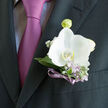 Pinned neatly above a mauve-lilac tie, this white orchid boutonniere looks like a moment of stillness amid the gentle excitement of a Clerkenwell wedding day. The orchid itself is full and open, its petals smooth and velvety, cupping a creamy centre that glows softly against the crisp charcoal lapel. There is no harsh white here; instead, a warm ivory undertone makes the bloom feel inviting and romantic. Surrounding it, light eucalyptus-style greenery and muted silvery leaves hug the stem, while tiny lilac blossoms and airy white gypsophila peep through, adding texture and a faint, powdery scent that calls to mind spring walks through Spa Fields or along Clerkenwell Green. A slim violet ribbon spirals around the bound stems, its satiny surface catching the light in small highlights that echo the sheen of the tie below. The ribbon's colour gently bridges the cool purple-pink tie and the delicate lilac accents woven into the design, making the composition feel harmoniously thought out rather than overly coordinated. Every element sits in perfect proportion: the flower neither too large nor too shy, the foliage framing but never overshadowing, the colours refined and modern. The way the boutonniere rests against the dark suit creates a sense of depth, as though the orchid is almost floating, lit by soft daylight from a nearby Clerkenwell townhouse window. It feels ready for a ceremony at a local registry office, a reception tucked away off Farringdon Road, or an intimate celebration in a hidden courtyard - a wearable expression of promise and quiet joy.