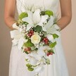 A bride in Clerkenwell, her white gown's lace catching hints of morning sunlight, is pictured from the torso down as she carefully holds a grand, waterfall-style bridal bouquet-a true showcase of a local florist's passion for opulent design. At the bouquet's crown, bold white lilies unfurl, each petal accentuated by soft reddish flecks that suggest fine artisan craftsmanship and evoke the gentle, natural beauty found in Clerkenwell's serene garden squares. Tucked amid these lilies, a central rose-petals tightly spun in creamy white-anchors the composition with timeless romance. Throughout the bouquet, lively tufts of chartreuse chrysanthemums and green pom flowers burst with contemporary cheer, mirroring the energy of the area's creative studios and galleries. Interspersed, clusters of shiny red berries introduce joyful contrast and an inviting touch of warmth, reminiscent of the festive displays in Exmouth Market. Delicate white phalaenopsis orchids trail down from the bouquet, their blossoms adding a graceful, cascading finish that draws the eye in gentle motion. Wispy sprays of baby's breath and fine grass-like greens provide soft framing and lightness, allowing each detail to breathe. The bride's gentle touch affirms the personal, bespoke nature of the floral design. Against the softly defocused background, this bouquet becomes the undisputed focal point-a fusion of classic wedding florals and lively local accents, perfect for ceremonies in the historic Clerkenwell area or for inspiring dreams of a perfect city celebration.