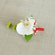 A meticulously crafted white orchid boutonniere is laid out on a softly textured, light beige linen background, the kind of neutral fabric you might see on a wedding table in a Clerkenwell townhouse venue. At the heart of the design, a single Phalaenopsis orchid bloom opens in pristine white, its broad petals smooth and almost porcelain-like, curving gently as if caught in a quiet breath. The throat of the orchid shows delicate shading, where soft yellow melts into subtle reddish-orange markings, drawing the eye into its intricate centre. Around this focal flower, airy clusters of white baby's breath are tucked in, their tiny blossoms forming a cloud-like halo that lightens the composition and adds a fine, lacy texture. To the left, two spherical lime-green chrysanthemum-style buds, tightly packed with spiky petals, provide a vivid modern contrast, their fresh colour echoing the energy of nearby Exmouth Market on a spring morning. Beneath the orchid, a broad, deep green leaf cradles the flowers, while a smaller dark green leaf peeks out near the stems, grounding the design in rich foliage. The stems themselves are neatly bound with a bold crimson satin ribbon, wrapped with careful precision and finished in a small, tidy knot that introduces a flash of celebratory red. The natural weave of the fabric beneath, visible in gentle, horizontal lines, emphasises the handmade, artisanal feel. Overall, the image captures a refined, contemporary boutonniere suitable for a groom or groomsman at a Clerkenwell wedding, radiating calm elegance and quiet joy.