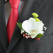 Pinned neatly to a classic dark lapel, this orchid boutonnière feels like a whispered promise of celebration on a Clerkenwell wedding day. The focal bloom is a pristine white orchid, its petals broad and smooth, almost translucent at the edges as if they were carved from polished ivory. A soft blush of buttery yellow glows from the flower's centre, drawing the eye inward with understated luxury. Just behind, two lime-green button chrysanthemums press forward, their tightly packed petals forming rounded domes that bring a spark of energy and fresh colour, much like the lively streets around Exmouth Market. Wisps of baby's breath unfurl delicately around the main flowers, each tiny white floret adding a feathery lightness, as though a veil of lace had been draped over the arrangement. Deep green leaves frame the flowers, glossy and neatly trimmed, giving quiet structure and depth. At the stem's base, a narrow ribbon of deep red is wound with precision, echoing the sheen of the matching tie and introducing a note of romance and warmth. Against the matte black suit, the white, green, and red palette appears crisp and refined, ideal for a groom or best man walking from a ceremony near St John's Square to an intimate reception nearby. Crafted with the eye of an experienced London florist, the piece balances texture, colour, and scale so it looks natural in photographs yet special up close. There is an imagined hint of clean, green fragrance from the chrysanthemums and a whisper of cool orchid freshness, creating an aura of calm confidence that carries the wearer from vows to evening toasts with effortless grace.