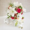 A romantic wedding-style arrangement in crisp white and rich red stands elegantly on a pristine white tabletop, framed by a softly textured beige backdrop that feels like draped fabric in a Clerkenwell loft venue. At the heart of the composition is a round, white container shaped like a basket, its open, criss-crossed weave adding a delicate, handcrafted look while keeping the design light and airy. Rising from this base, several large oriental lilies in glowing white open fully, their petals smooth and sculptural, with warm-toned stamens that lend a natural, slightly wild charm despite the arrangement's careful styling. Curled among the lilies are velvety red roses, their deep, saturated hue and tightly layered petals suggesting passionate toasts and late-night stories around the wedding table. Fine sprigs of white baby's breath float between the larger blooms, softening the strong palette, while small, bright red hypericum berries and scattered bright green foliage bring a touch of woodland detail, as if gathered on a walk through a nearby London garden. On the right-hand side, a cascade of white phalaenopsis orchids spills gracefully down from the main bouquet, their blooms arranged along a gentle arching stem, each flower marked with subtle yellow and blush tones at the centre. To the left, three white woven spheres lie on the tabletop, echoing the basket's texture and helping to frame the scene. The overall effect is one of balance and understated luxury, perfect for a Clerkenwell wedding reception, an elegant anniversary dinner, or any special event that calls for a sophisticated floral focal point.
