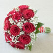 A hand-crafted bouquet of romantic red roses and airy white baby's breath is shown in a calm, diffused daylight that feels very much like a Clerkenwell studio with tall sash windows. The roses are a deep, velvety crimson, each blossom opening in layered swirls that suggest luxury and heartfelt passion. They are nestled closely together in a rounded, dome-like shape, creating a sense of fullness and generosity, while fine sprays of gypsophila weave between them like soft lace, bringing lightness and gentle movement to the design. Subtle touches of rich green foliage frame the flowers, their smooth leaves giving a cool, refreshing edge that enhances the intensity of the reds and whites. The stems are neatly gathered and wrapped in elegant ribbon, finished with tiny pearl accents that call to mind bridal jewellery and timeless ceremonies in nearby Clerkenwell churches. The bouquet carries an almost tangible scent in the imagination: the sweet, classic aroma of fresh roses, softened by the clean, delicate character of the gypsophila. Its mood is one of quiet grandeur and intimate celebration, equally at home in a bridal suite overlooking Farringdon Road or in the hands of someone arriving at a registry office near Spa Fields. Every detail-from the precise placement of each rose to the airy spacing of the baby's breath-speaks of an experienced florist's touch and a deep understanding of how flowers can embody love, commitment, and the special atmosphere of a wedding day in Clerkenwell.