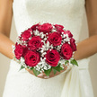 In this image, a bride holds a perfectly rounded bouquet of red roses and white baby's breath, the flowers resting softly against a pale, textured wedding dress. The roses are a deep, classic red, each bloom at its prime, with unfurling petals that form luxurious, velvet-like rosettes. They sit tightly gathered at the centre, creating a sense of richness and warmth, like a glowing ember of colour amid the gentle tones of the gown. Surrounding them, sprays of white gypsophila create a delicate, cloud-like border, their tiny blossoms catching the light and adding an airy, romantic shimmer. The stems are bound together neatly, likely wrapped in ribbon to sit comfortably in the bride's hands as she walks through Clerkenwell's quiet lanes, perhaps heading from a townhouse near Spa Fields to a nearby venue. The bouquet feels at once traditional and intimate, reflecting the kind of thoughtful design a local florist might prepare for couples marrying in historic churches around Clerkenwell Green or in stylish converted warehouses by Farringdon. Though you cannot smell it, the photograph suggests a soft blend of rose perfume and fresh foliage, a scent that would linger gently throughout the ceremony. The composition is calm and balanced, with the round form of the bouquet mirroring the sense of completeness and unity that a wedding day brings, making it a fitting floral companion for vows exchanged in this characterful corner of London.