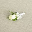 A finely detailed photograph shows a single white rose boutonnière placed diagonally across a softly textured, light beige fabric that looks like natural linen, the gentle crosshatch of the weave clearly visible. At the heart of the design sits a creamy ivory rosebud, its velvety petals still mostly furled, with just the outer edges beginning to unfurl in a quiet promise of bloom. The rose's base is cradled by faint green sepals, which merge into a cluster of slender, deep green leaves fanning out behind the flower. To one side of the rose, airy white baby's breath unfurls in tiny cloud-like blossoms, each minuscule flower adding a misty softness and a sense of lightness to the compact arrangement. Threaded among these blooms, a few finer, fern-like sprigs introduce delicate texture, suggesting the careful hand of an experienced florist used to creating wedding pieces for intimate ceremonies in Clerkenwell's townhouses and registry offices. The stems are tightly and neatly bound in a pure white satin ribbon, its surface smooth and faintly luminous in the soft, diffused light. Just below the bloom, the ribbon is finished with a small, perfectly tied bow, giving the boutonnière a polished, ready-to-pin appearance, ideal for the lapel of a groom or groomsman arriving from Farringdon Station to a nearby venue. Shadows are gentle and understated, allowing the creamy whites and fresh greens to appear calm and timeless. The overall mood is one of classic elegance and quiet romance, making this boutonnière a graceful accent for weddings, civil partnerships, or formal celebrations around Clerkenwell and central London.