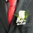 A refined boutonniere sits neatly on the lapel of a deep charcoal suit, its white rose the clear focal point against the dark cloth, as if crafted for a wedding just off Clerkenwell Green. The rose is a creamy, luminous white, with petals that curve in soft, natural layers, neither tightly closed nor fully blown, suggesting a moment of perfect readiness. Surrounding its base are clusters of snow-white baby's breath, their minuscule blooms forming a delicate halo that lightens the overall composition. Wisps of fine greenery and slender fern fronds arch outward and downward, introducing movement and a touch of garden-like spontaneity that contrasts beautifully with the structured suit. At the stem's base, a small white satin bow has been tied with careful hands, the ribbon smooth and slightly glossy, echoing the formality of crisp shirts and polished shoes. The fabric of the suit reads as well-tailored and subtly textured, framing the floral work without stealing attention. The scene suggests an indoor space filled with gentle anticipation-a registry office near Farringdon, perhaps, or a chic Clerkenwell warehouse venue preparing for vows. The boutonniere's design feels timeless and thoughtful, suited to a groom, best man, or honoured guest who values understated style. There is an air of polished restraint, yet also quiet emotion, as if each petal carries a story of promises and shared memories. The combination of soft white blooms, fresh greenery, and careful finishing details reflects the expertise of a local florist who understands how a small floral accent can define the character of an entire celebration.