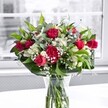 A vibrant, hand-crafted bouquet of red roses, red carnations, white alstroemeria, and airy baby's breath stands proudly in a clear glass vase on a bright white tabletop, bathed in soft natural light from a nearby window in Clerkenwell. The rich crimson roses are the stars of the arrangement, some fully open with velvety petals spiralling outward, others still in tight buds that promise to unfurl over the coming days. Nestled beside them, ruffled carnations in the same deep, romantic red add fullness and an old-fashioned charm, their many layers catching the gentle daylight. Threaded between the bold reds, white alstroemeria - some still in green-tipped bud, others partially open - reveal their slender, trumpet-shaped petals, bringing a quiet, graceful contrast. Fine clouds of delicate white gypsophila dance through the bouquet, softening the colour blocks and giving the impression of tiny stars scattered across a night sky. Around the base, broad, glossy green leaves and more slender, lighter foliage cradle the flowers, creating a natural collar that frames the central blooms and adds a lush, fresh backdrop. Through the crystal-clear vase, neatly trimmed stems are visible, immersed in clean water that reflects the surrounding light, emphasising the bouquet's freshness as if it has just been arranged in a local Clerkenwell studio. Behind, the window view is intentionally blurred and almost monochrome, making the reds, whites, and greens appear even more vivid. The overall mood is one of joyful elegance, perfect for a romantic anniversary, an intimate dinner near Exmouth Market, or a heartfelt thank-you delivery to an office off Farringdon Road.