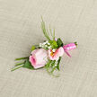 This boutonniere presents a poised and graceful composition, centred on a softly blushing pink rose whose petals curl outwards in delicate, velvety layers. The colour shifts from a pale, almost porcelain pink at the tips to a fuller rose hue near the heart, giving the flower a luminous, gentle glow, like the last of the evening light over Clerkenwell's brick terraces. Around it, pastel alstroemeria blooms offer a subtle contrast: their petals are lightly brushed with blush and cream, each bloom finished with a tiny splash of yellow at the throat, as though someone has painted them with the finest of brushes. Threaded between these main flowers, airy sprigs of white gypsophila provide a cloud-like softness, creating a halo that makes the rose appear to float. Slender green fronds and curled leaves peek out from behind, adding a crisp, refreshing note and a suggestion of movement, reminiscent of foliage stirred by a breeze along Farringdon Road. The stems are drawn neatly together and wrapped in a gleaming pink satin ribbon, its smooth surface catching subtle highlights, keeping everything tidy and ready to pin to a jacket. Resting on textured fabric-perhaps a suit laid out before a ceremony-the arrangement looks both intentional and effortless, crafted with care for a special day. You can almost imagine the cool touch of the ribbon, the velvet of the rose, and the faint, uplifting scent that lingers as the wearer walks into a wedding, civil ceremony, or celebration in one of Clerkenwell's historic venues, feeling quietly polished and thoughtfully adorned.