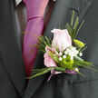 The photograph captures a romantic wedding boutonnière in refined detail, as if it has just been adjusted on a groom's lapel outside a Clerkenwell townhouse. At the centre is a blush-pink rose, its petals layered in soft spirals, shading from near-white at the outer edges to a tender, powdery pink towards the heart. The rose feels full of promise, not fully open yet clearly alive with potential, like the beginning of vows spoken at the local town hall. Surrounding the bloom, neat greenery in cool, fresh tones provides a sculpted frame, the leaves smooth and slightly glossy, offering a clean contrast to the rose's velvety texture. Threaded through the foliage is a slim stem of white gypsophila, the tiny flowers forming a delicate constellation of cloud-like dots, evoking a lightness and sweetness that never overwhelms. Tucked in close is a green button chrysanthemum, round and compact, its many tiny petals tightly packed to create a crisp, modern accent that suggests contemporary floral design so often favoured by couples marrying around St John's Square. A single elongated leaf arcs gently, directing the eye downward to where a narrow lavender-mauve ribbon embraces the stems. The ribbon is tied neatly with its ends trailing just enough to feel festive without fuss, hinting at satin lapels and polished shoes. The entire boutonnière is bathed in soft, even light, showing every subtle vein in the leaves and every curve of the rose. It radiates quiet elegance and personal care, the sort of handcrafted finishing touch a local Clerkenwell florist would prepare for a wedding or formal celebration that values intimacy, artistry, and a sense of place.