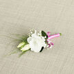 A delicately crafted boutonniere lies against a light, textured fabric background, evoking the understated elegance often seen in Clerkenwell's converted warehouses and studio spaces. The focal point is a generous white lisianthus bloom, open and ruffled, its creamy petals layered in soft curves that suggest both luxury and gentleness. Just beside it, two lisianthus buds nestle close, one still tightly closed and the other beginning to unfurl, like a visual whisper of love in its early chapters, ready to be celebrated at a wedding near Farringdon or an intimate gathering off Clerkenwell Road. Fine sprigs of baby's breath weave between the blooms, their tiny white flowers forming a delicate constellation that softens the composition and gives it a dreamy, mist-like quality. Slender green leaves and wispy foliage extend from the base, adding a sense of movement, as though a light breeze might stir them in a hidden courtyard garden behind a local townhouse. The stems are gathered with care and wrapped in a vivid pink satin ribbon, smooth and gleaming, which winds tightly around the base before forming a small, tidy bow. This ribbon introduces a cheerful pop of colour that lifts the design without overpowering the calm, white-and-green palette. The boutonniere feels handcrafted and considered, the sort of piece a local florist would prepare on a quiet morning before delivering it across Clerkenwell for a city wedding, civil ceremony, or stylish rooftop celebration, bringing a little moment of romance to the wearer's lapel.