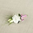 A delicately crafted wedding boutonnière rests at a gentle diagonal on a softly textured light beige fabric, the weave reminiscent of fine linen you might find in a Clerkenwell design studio. At its heart is a single, fully opened white lisianthus bloom, its ruffled petals layered like soft silk, forming a rounded cup of creamy white that feels calm and refined. Two slender lisianthus buds, tinged with pale green and cream, nestle just to the left and below, suggesting flowers that will open later in the day, perhaps just in time for evening photographs along Clerkenwell Road. Tiny sprays of white gypsophila are threaded through the composition, their airy, cloud-like clusters adding a sense of lightness and quiet romance. Slender, grass-like blades of fresh green foliage sweep outward towards the lower left, creating graceful movement, while a broader, darker green leaf sits behind the main bloom, giving depth and structure. The stems are neatly gathered and wrapped at the base in a vibrant fuchsia satin ribbon, tied into a small, jaunty bow that adds a playful, modern splash of colour against the neutral background. The satin catches the soft, even light, giving a subtle sheen that hints at celebration. The overall mood is intimate and elegant, the sort of boutonnière you could imagine pinned to a groom's lapel for a ceremony in a hidden Clerkenwell courtyard, captured in close-up to highlight every petal, texture, and thoughtfully placed detail of this small but meaningful floral accent.
