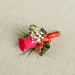 A handcrafted boutonniere is laid gently on a softly textured, light beige linen background, the kind of woven fabric you might find in a Clerkenwell townhouse overlooking Exmouth Market. At its heart sits a single deep fuchsia rose, petals velvety and tightly layered, spiralling outward in a rich swirl of hot pink that suggests romance, excitement, and the promise of celebration. Nestled around the rose are airy clusters of tiny white blooms, unmistakably baby's breath, their cloud-like softness catching the light and adding a sense of innocence and delicacy. Between these white blossoms shine small, glossy red berries, likely hypericum, each one rounded and plump, giving a cheerful, almost festive sparkle of scarlet that plays beautifully against the rose. Surrounding the flowers is a collar of lush green foliage: some leaves broad and dark, carefully curled to frame the main flower, others fine and feathery, like light asparagus fern, stretching outward to create a sense of movement and natural grace. The stems are bound tightly and neatly in a vivid red satin ribbon, wrapped several times to form a smooth, luxurious handle, finished with a tidy knot or tiny bow that adds polish and quiet sophistication. Soft, even daylight glides across the scene, revealing every subtle texture-the rough weave of the fabric, the gentle sheen of the ribbon, the glossy surface of each berry. The overall mood is one of refined joy and understated glamour, suitable for a groom or groomsman stepping out of a registry office near Clerkenwell Green or arriving at a chic city wedding on St John Street, wearing this small but meaningful floral detail on the lapel as a symbol of love, celebration, and carefully considered style.