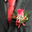 A detailed, high-angle close-up shows a classic wedding boutonniere pinned neatly to the left lapel of a dark charcoal grey suit jacket, the kind you might see on a groom stepping out of a townhouse near Exmouth Market in Clerkenwell. At the heart of the buttonhole sits a single deep red rose bud, tightly furled yet full of promise, its velvety petals catching the soft indoor light and drawing the eye immediately. Around the base of this rose, clusters of tiny white gypsophila create a cloud-like halo, their airy texture gently brightening the arrangement and softening the strong red tones. Nestled between the flowers are several small, glossy red berries, perfectly polished, mirroring the richness of the rose while adding a subtle, tactile contrast. Slim, grass-like fronds of greenery arc gracefully from behind, fanning outward with a slightly wild, natural feel, while broader, darker green leaves are wrapped close to the stem, giving structure and a sense of careful craftsmanship. A narrow red satin ribbon is tied into a compact bow at the base, its sheen echoing the luxurious fabric of the bright red tie visible just beneath the lapel. The tie is knotted with precision, its bold colour reinforcing the romantic red palette and framing a discreetly patterned light grey dress shirt, whose fine checks or houndstooth weave add quiet sophistication. The overall scene evokes a poised, celebratory moment, perfectly suited to a Clerkenwell wedding, civil ceremony at the Town Hall, or elegant evening reception, and reflects the thoughtful attention to detail that elevates men's formal wear for a truly memorable occasion.