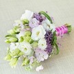 Resting on a creamy, finely woven linen backdrop as soft as the morning light spilling through Clerkenwell's quiet backstreets, this radiant hand-tied bouquet showcases the expertise and artistry that define local floral design. At its core, generous white Lisianthus (Eustoma) blossoms unfurl with ruffled precision, their centers softly yellow, surrounded by delicate, lime-green and white buds suggesting the gentle promise of spring. Threads of pastel lavender-pink stock and sprays of deep purple allium stars sweep through the composition, adding tensile contrast and airy sophistication that echo the vibrant creativity found in the heart of EC1. Elegant strands of arching bear grass and flecked foliage nestle amongst the blooms, lending a sense of movement and lively detail reminiscent of Clerkenwell's pocket gardens. Bound with a striking fuchsia satin ribbon and an artful tangle of glossy white pearls, the bouquet's stems convey luxury and personal flair. This thoughtfully crafted arrangement feels at home in a modern wedding-or any celebration-near St John's Square or Clerkenwell Road, radiating romance and freshness. Its palette, scent, and textures encapsulate both joyous exuberance and delicate serenity, making it the perfect visual for stories about springtime weddings, bridesmaid bouquets, or sophisticated event florals in this distinctive London neighbourhood.