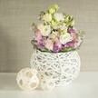 In softly diffused daylight characteristic of Clerkenwell flats, a handcrafted floral display brims with quiet charm and texture. At centre stage, velvety eustoma blooms in shades of milky white and pale yellow pool together in ruffled, layered clusters, exuding subtle sophistication. Tall stems of pink matthiola stock elegantly punctuate the bouquet, their pastel hues introducing delicate height and whimsical beauty. Underneath, a tapestry of vivid purple filler flowers lines the base, infusing depth and evoking the artistic spirit found on Clerkenwell's vibrant streets and community gardens. Fresh deep green foliage sweeps around the edge, its gentle contrast bringing the pastel palette into sharper focus. The flowers are anchored in a unique white vase, spherical and woven from painted rattan in an open, lacy design-an echo of local creative makers and modern design studios. Adjacent to the main arrangement, two light wood decorative spheres, with airy open-weave sides, rest on a pristine tabletop, continuing the scene's artisanal and natural theme. A soft beige wall with subtle, fabric-like texture forms the background, ensuring the bouquet's hues and details remain centre stage. Every element-inspired by Clerkenwell's dynamic mix of heritage and innovation-speaks of fresh beginnings and sophisticated simplicity, ideal for marking life's special moments or styling contemporary local interiors.