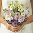 This image shows an elegant, hand-crafted bouquet that feels like a piece of living poetry, the kind of bridal flowers you might see being carried from a townhouse near St John Street towards a ceremony in Clerkenwell. At the heart of the design, creamy white lisianthus and soft blush stock blooms unfurl in gentle layers, their petals almost translucent where the natural light brushes across them. Hints of pastel lilac weave through the whites, leading the eye down towards a base of rounded, starburst-shaped purple allium heads, each tiny floret adding intricate detail and a sense of depth. The bouquet is gathered in a relaxed yet deliberate spiral, creating a light, open dome at the top where each flower has space to breathe, while the stems nestle closely together at the bottom, forming a neat, tidy handle that would feel cool and reassuring in the hand. Encircling the flowers, fine strands of fresh green bear grass loop and arc like delicate ribbons, structured yet fluid, lending the bouquet a modern, almost architectural twist without losing its romantic softness. The interplay of creamy whites, tender lilacs, and vivid greens suggests freshness and new beginnings, as though the bouquet has just arrived from an early-morning floral delivery to a Clerkenwell studio. You can almost sense the subtle fragrance-a sweet hint from the stock, a clean greenness from the foliage-rising gently rather than overwhelming the senses. This arrangement feels perfect for weddings, engagements, or vow renewals, its understated elegance and thoughtful craftsmanship turning a handful of blooms into a keepsake moment that lives in memory long after the petals fade.