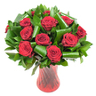 A lavish, rounded bouquet of roughly a dozen deep crimson roses fills the frame, each bloom opened into a perfect spiral of velvety petals whose colour reads almost wine-dark at the heart and brightens toward the petal edges. The roses sit closely clustered to form a dense, domed profile; soft, even studio light skims the petals, revealing fine veining and a subtle satin sheen that suggests freshly cut stems. Nestled between the heads is an abundance of dark green foliage - glossy, flat leaves like Ruscus or Salal form a rich, verdant backdrop while broader, emerald Aspidistra-style leaves are expertly curled and rolled into sculptural loops and tubes. Those folded leaves create rhythmic arcs that cradle each rose and add a contemporary architectural counterpoint to the classic blooms. The arrangement is anchored in a tall, slender translucent ruby-red glass vase with a faintly ribbed, twisted texture that catches the light in vertical highlights, echoing the roses' passionate tone. Set against a clean white background, the strong reds and deep greens pop with clarity; the composition reads as both intimate and formal, a floral statement that radiates romance and refinement. Imagine the faint, warm scent of rose and green leaves, and the way this piece would command a dining-table centre or an anniversary gift delivered across Clerkenwell. The bouquet's balance of traditional red roses and modern foliage styling makes it equally suited to Valentine's Day, an elegant proposal, or a quiet, heartfelt celebration in a nearby courtyard off Exmouth Market.