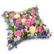 In this photograph, a luxurious pillow-shaped floral tribute is shown from above, its soft rectangular form neatly defined yet gently rounded at the corners, like a cushion laid down in quiet memory. The entire surface is carpeted with flowers, dominated by roses in shades of vivid magenta-fuchsia, soft pink, and creamy pale yellow, many of them fully unfurled so that each petal can be distinguished. Between the roses, rich royal purple lisianthus blooms provide a velvety contrast, their darker tone adding a sense of depth and solemn grace. Tiny lavender-hued filler flowers, reminiscent of limonium or statice, along with clusters of blush pink spray roses, weave through the composition, filling every space and lending a fine, lace-like detail to the overall look. The blooms are supported and framed by layered greenery: rounded, silvery eucalyptus leaves bring a cool, misty green, while darker, fern-like foliage adds structure and texture, making the cushion appear soft yet reassuringly full. Set against a plain white background with a faint shadow beneath, the tribute stands out clearly, as if ready to be carried from a Clerkenwell florist's workroom to a service near Charterhouse Square or a quiet goodbye at a local crematorium. The colours and shapes together create a mood of tenderness and respect, making this floral cushion a fitting choice for families seeking a compassionate, beautifully made sympathy arrangement in the heart of Clerkenwell.