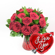 A refined yet passionate composition of roses and symbolism is set crisply against a stark white background, capturing the essence of a grand romantic gesture in Clerkenwell. At the centre, an opulent cluster of deep red roses rises from a ruby-tinted glass vase, each bloom carefully placed to create a rounded, abundant shape. There are roughly a dozen or more roses, some petals still cupped and others unfolding into broad, velvety spirals, giving the bouquet a sense of movement and life. Fresh green foliage is threaded throughout, its leaves a glossy, saturated green that frames and supports the flowers, suggesting the attentive conditioning work of a professional florist. The vase itself is a clear glass form with a ruby-red hue and subtle fluted ridges that run vertically, catching points of light and giving the illusion of gentle ripples. In the foreground to the right, a bright, metallic red heart-shaped foil balloon leans slightly toward the arrangement, partly overlapping the vase and blooms. Its surface is mirror-smooth and reflective, and across the middle, in flowing white cursive, are the words I Love You, easily legible even from a distance. A stylised silver arrow, echoing Cupid's, appears to pierce the heart from lower left to upper right, adding a whimsical, storybook charm. With the neutral background eliminating all distractions, the bold reds and greens stand out in vivid contrast, making this image a perfect visual for expressing love, whether for a Valentine's Day surprise near Clerkenwell Green, an anniversary toast at a local restaurant, or a spontaneous romantic delivery to someone special.