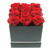 The image reveals a beautifully composed square rose box that feels like a natural fit for the stylish interiors around Clerkenwell. At the centre is a matte black cube-shaped box, its surfaces smooth and untextured, with sharp edges that lend the arrangement a contemporary, almost gallery-like presence. Around the lower section of the box, a thin white stripe runs in a clean horizontal band, offering a subtle contrast much like the refined details found in Clerkenwell's modern cafés and design studios. Rising from the open top are twelve perfectly presented red roses, aligned in a precise three-by-four grid so that each blossom is nestled close to its neighbours. The roses are fully open, their petals thick and velvety, spiralling outward in intricate layers that create small pools of shadow at the heart of each bloom. Their rich ruby colour is intensely saturated, with deeper crimson tones tucked into the inner whorls and slightly lighter edges where the soft studio lighting brushes across them. The flowers just peek above the rim of the box, forming a plush, cushion-like surface that looks both structured and indulgent. The white background is completely uncluttered, ensuring that all attention is drawn to the interplay between the dark box and the vivid roses. Gentle, even lighting smooths away harsh contrasts, instead creating subtle shading along the petals and at the corners of the container to suggest volume and depth. The overall mood is polished and romantic, making it ideal for a Valentine's Day delivery to a flat near St John's Square, an anniversary surprise at a restaurant off Charterhouse Street, or a special gift marking an engagement celebrated in one of Clerkenwell's converted warehouse lofts.