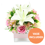 A graceful, dome-shaped bouquet of fresh pink and white flowers is shown against a bright, uncluttered white background, as though ready to leave a Clerkenwell florist's counter for hand delivery. At the centre, two generously open white Oriental lilies dominate the eye, their silky petals spreading outward and curling delicately at the tips, exposing long, dark reddish-brown stamens that add dramatic depth to the otherwise soft palette. Encircling these lilies is a profusion of blush and mid-pink roses, some still cupped tightly like miniature sculptures, others unfurling into full, layered blooms that look plush and romantic. Threaded between the roses and lilies are clusters of alstroemeria in shades of pink and white, with several petals marked by gentle yellow washes and tiny speckles that bring a playful, intricate detail to the arrangement. Fine sprays of white baby's breath are sprinkled throughout, giving a light, airy lift to the design, almost as if the flowers were floating on a cushion of cloud. A selection of lush greenery - broad, glossy leaves, softer fronds, and slender, arching blades - frames the blooms and reinforces the sense of natural abundance, reminiscent of leafy corners around Clerkenwell Green or the quiet side streets near St John Street. The entire composition is set neatly in a low, square clear glass vase, its see-through sides revealing the carefully trimmed stems and fresh water inside. A pale pink satin ribbon wraps horizontally around the vase's middle, forming a smooth band that adds a gentle, gift-like finish. In the bottom right corner, a bright orange circle with bold white lettering clearly reads VASE INCLUDED, making it easy to understand that the container accompanies the flowers. The image conveys a feeling of calm celebration, ideal for marking special occasions, saying thank you, or sending a little everyday luxury to someone in Clerkenwell.